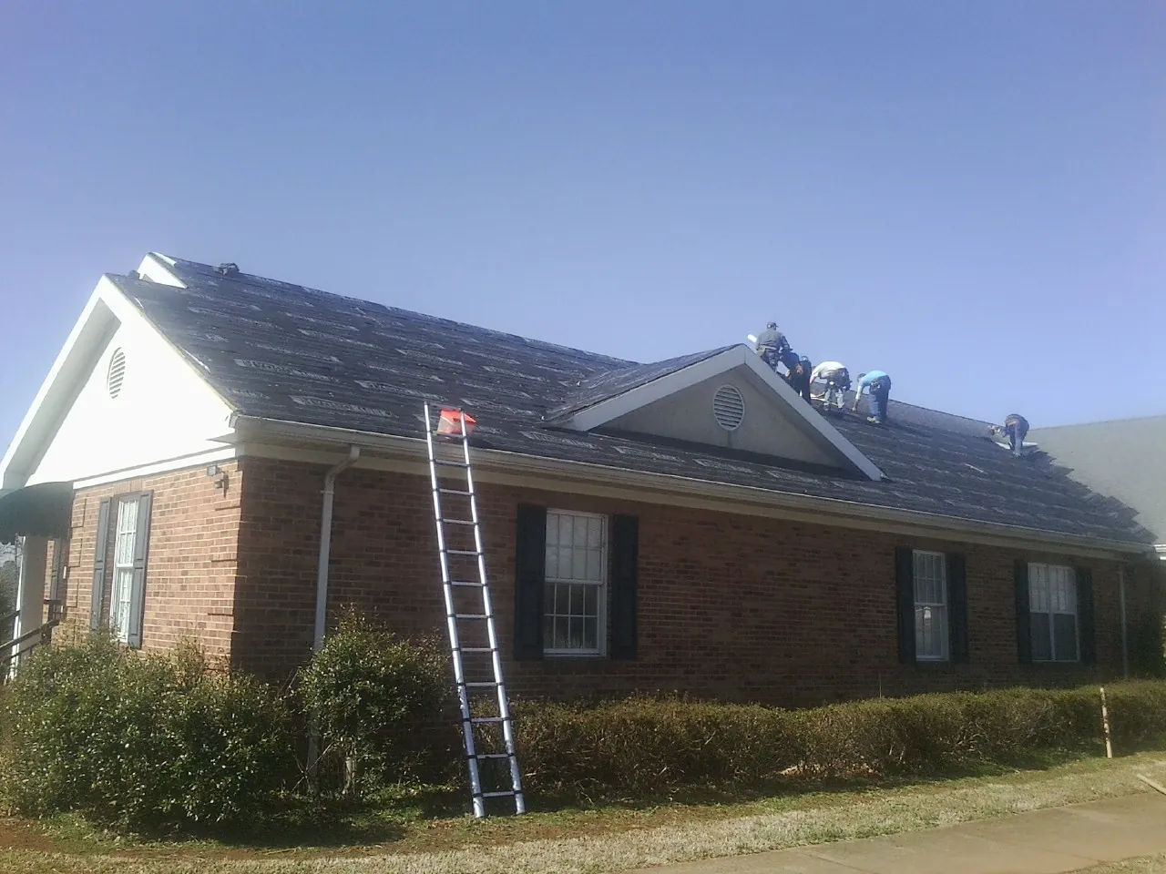 Crew tearing shingles off a brick ranch roof with a ladder set — Metal Roof Repair work by our crew in Knowlesville
