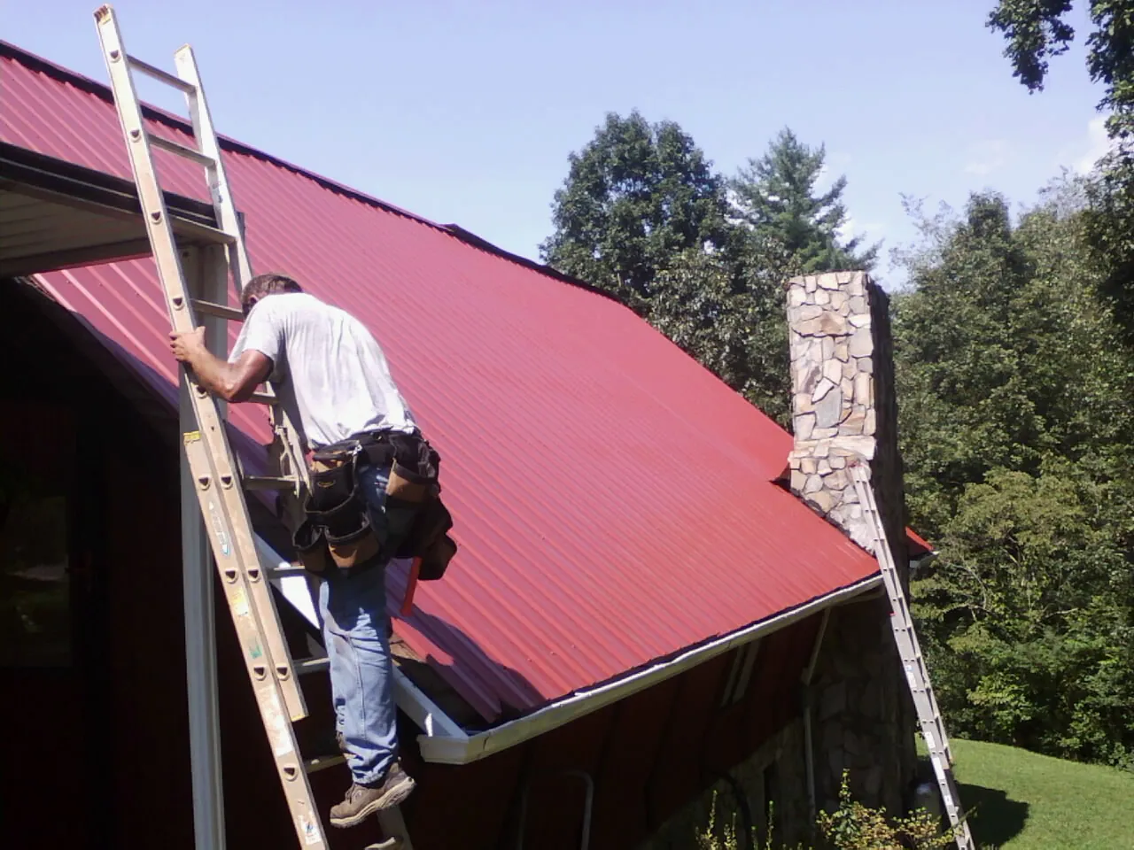 Roofer on a ladder beside a red metal roof and stone chimney — Soffit & Fascia Repair by Roofing Brothers in Knowlesville