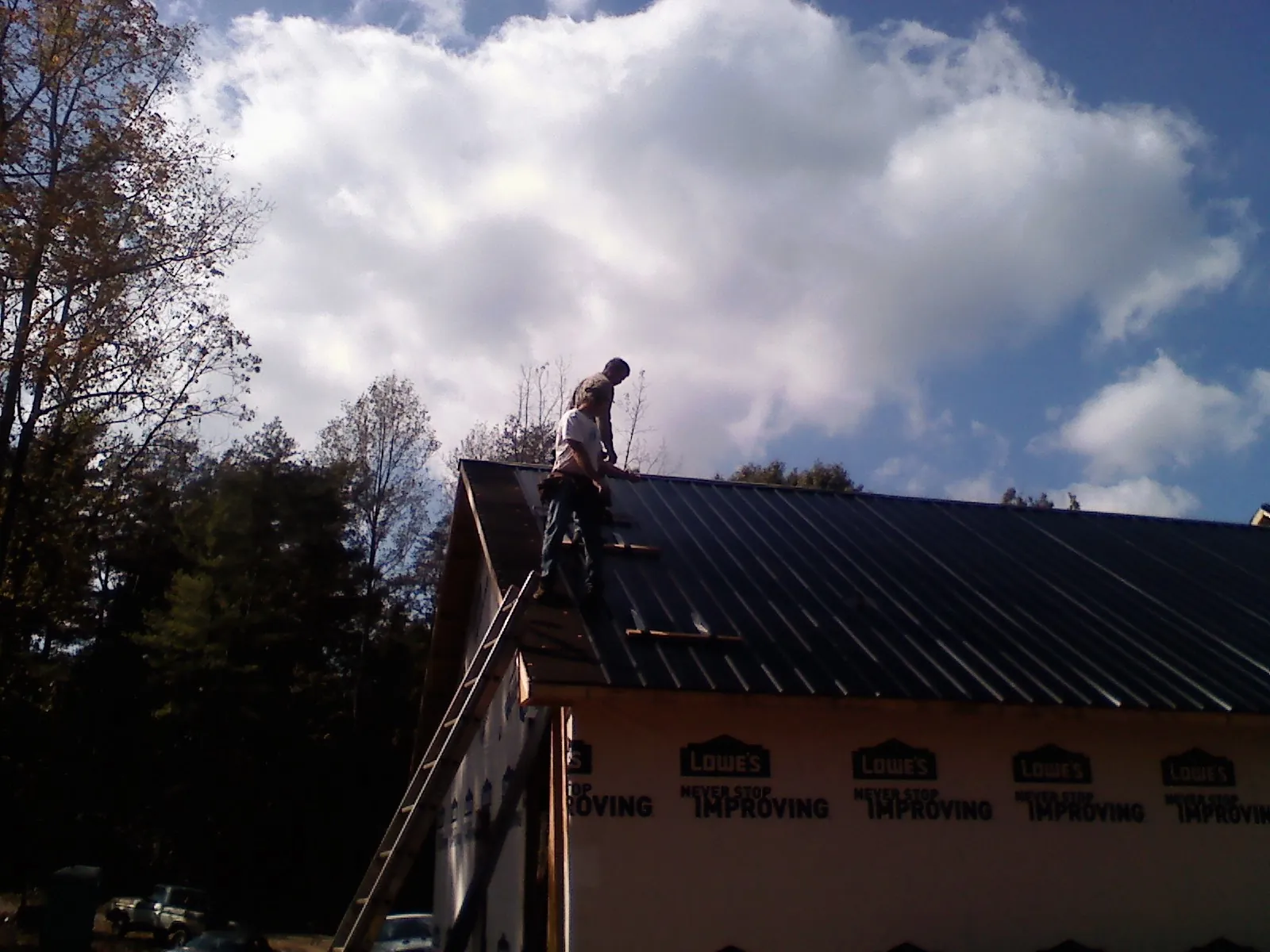 Roofer fastening dark metal panels high against a cloudy sky, from a Roof Inspection job in Knowlesville
